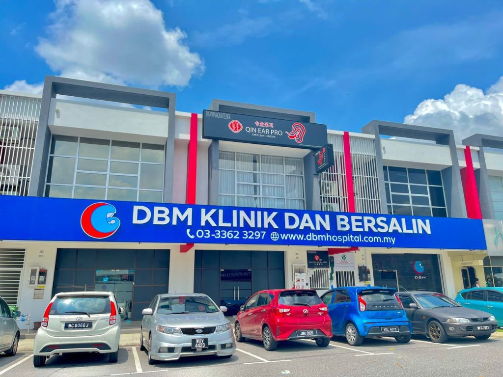 Front view of DBM Klinik dan Bersalin Bukit Raja showing the clinic sign with the logo, contact number, and website address. The sign reads 'DBM KLINIK DAN BERSALIN' with the website 'www.dbmhospital.com.my' and phone number '03-3362 3297'. The building facade is modern with large windows and a blue sign. Several cars are parked in front of the clinic