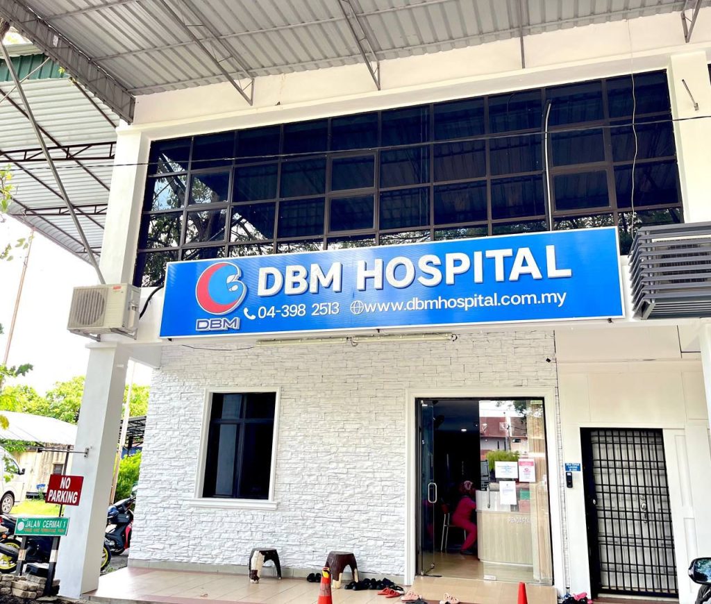 Front entrance of DBM Hospital Permatang Pauh showing the hospital sign with the logo, contact number, and website address. The sign reads 'DBM HOSPITAL' with the website 'www.dbmhospital.com.my' and phone number '04-398 2513'. The entrance area has a white brick facade, a glass door, and a window.